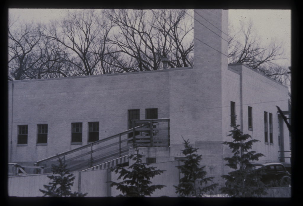 The original meat lab constructed at UW Madison was a one story limestone colored brick building with a chimney on one corner. The building has many trees around it and a period car is parked outside.