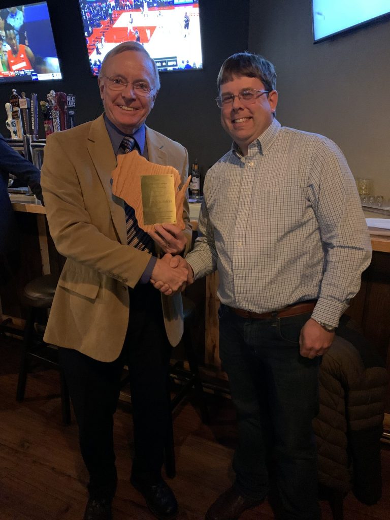Dan Schaefer holds a plaque and shakes a man’s hand as the two pose for a photo at a restaurant bar.