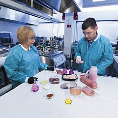 Two researchers wearing safety glasses and blue lab coats stand around a table with various meat products and several glass containers holding different colored liquids.