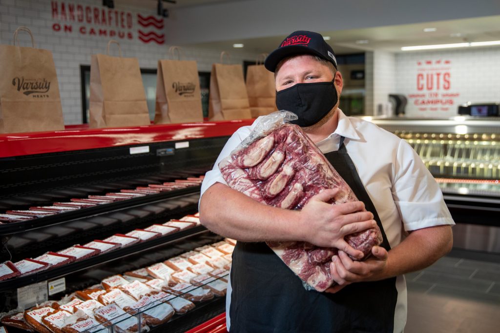 Mitch Monson holds a vacuum wrapped meat product in front of a display case in Bucky’s Varsity Meats.