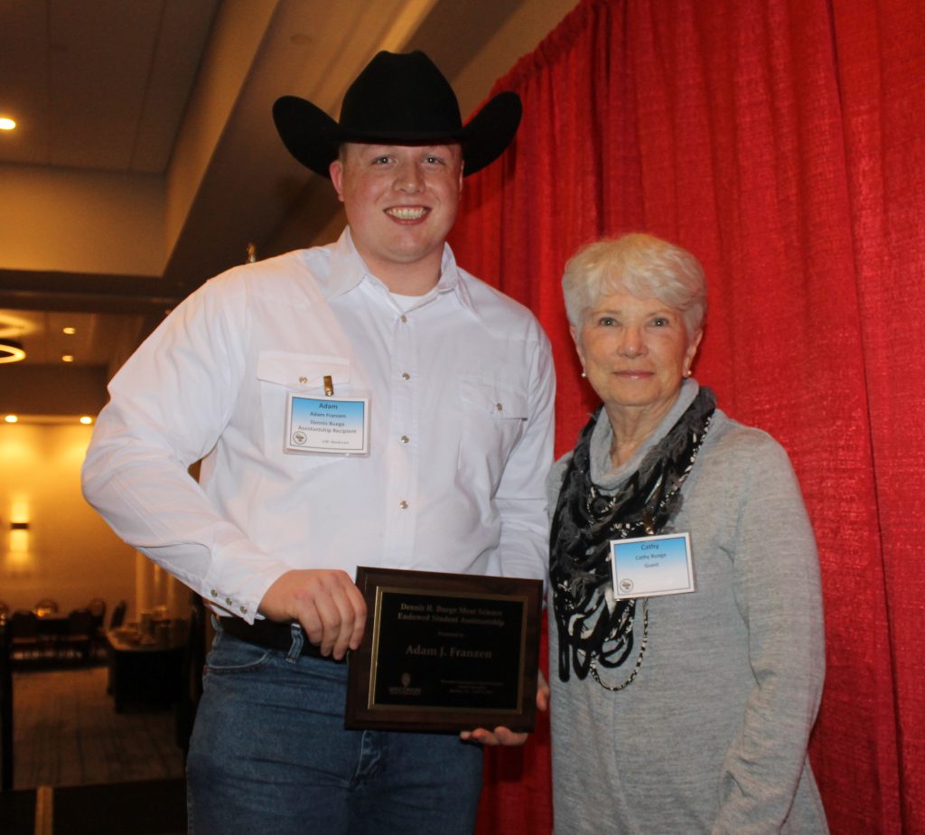 Adam Franzen, wearing a cowboy hat, holds a plaque presented by Cathy Buege while standing beside her in a hotel banquet hall.