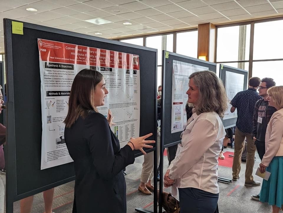 Ashley Tarcin gestures with her hands as she explains her research to a woman listening while the two stand in front of an easel holding Ashley's award winning poster.