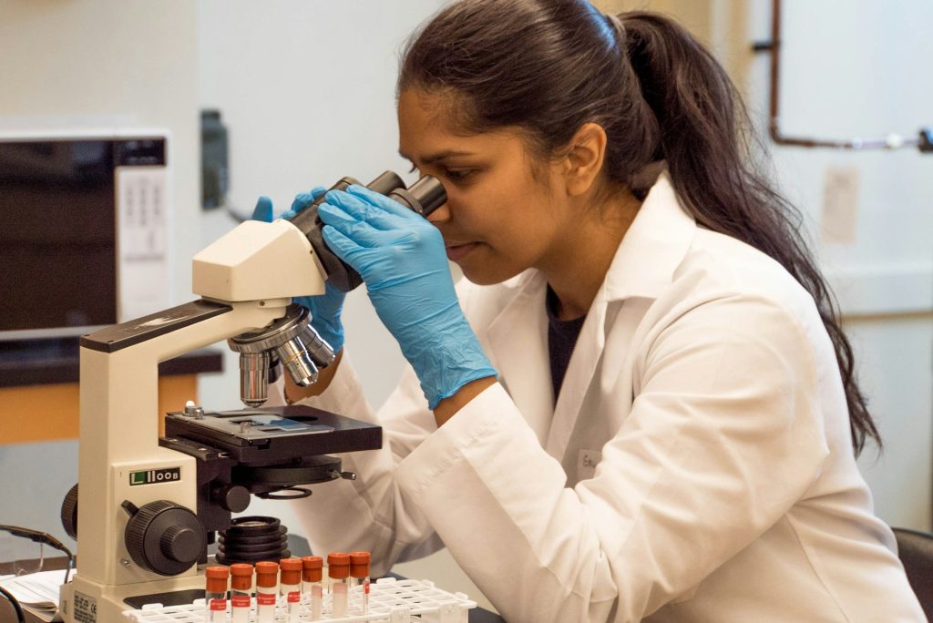 A woman in a lab coat views a sample through a microscope.